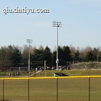 A sports field with a group of people engaging in athletic activities in the background. There is sports equipment like a soccer goal and a scoreboard visible. A chain-link fence runs across the foreground, and a statue or sculpture resembling an eagle is also seen. Trees and tall floodlights are positioned behind the field.